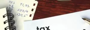 A document labeled "tax deductions" lies on a wooden desk next to a pen, a notebook, glasses, and a piece of paper with handwritten numbers.